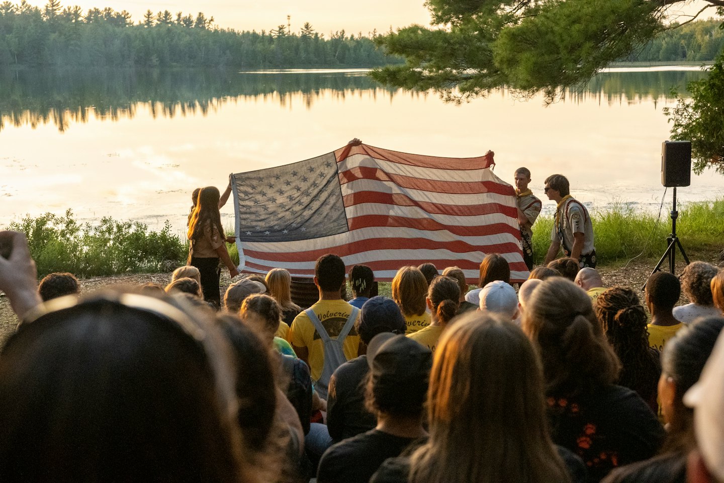 Scouter håller amerikansk flagga vid sjön i solnedgången med skog.